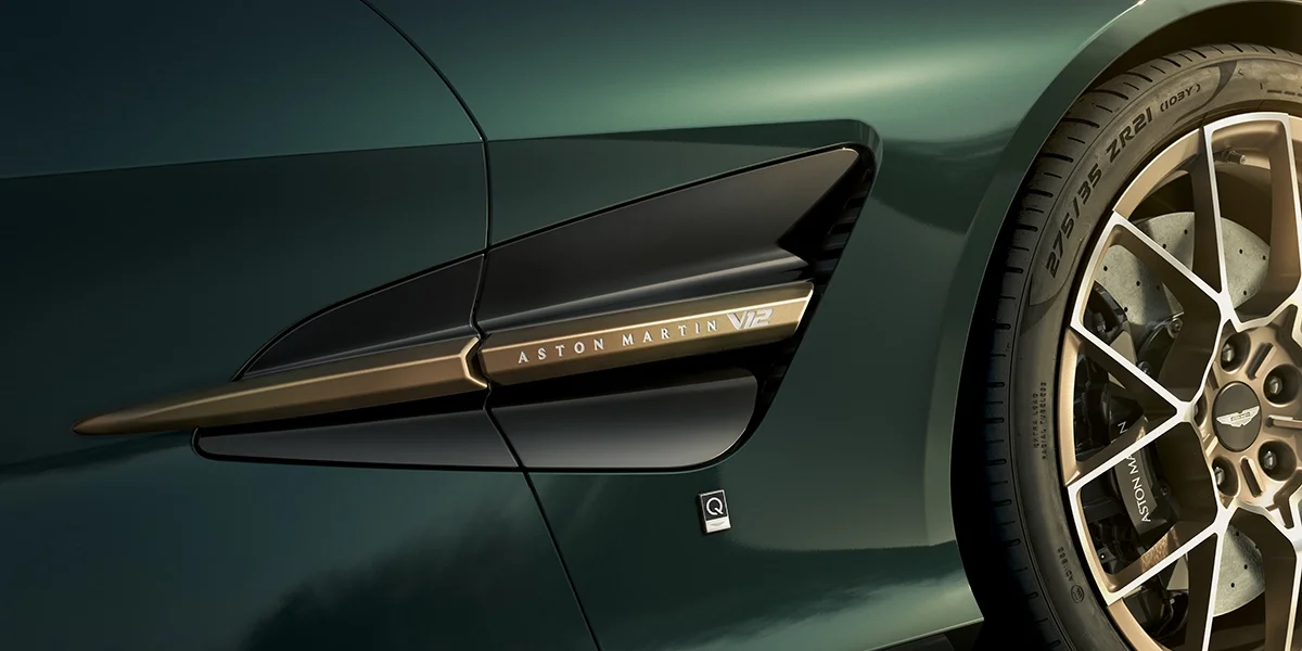 Detail of Aston Martin Vanquish Volante center console featuring gold engine start button, luxury wood finish, and precision-milled controls.