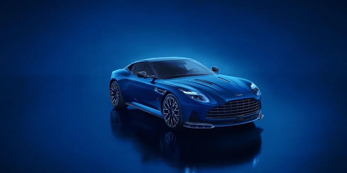 Front close-up of the Aston Martin DB12 S highlighting its signature grille, sleek blue paint finish, and illuminated LED headlights against a dark blue background.